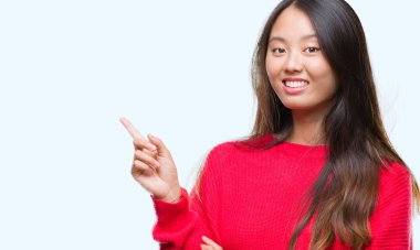 Kameraya bakarak yan el ve parmak ile işaret yüzünde kocaman bir gülümseme ile izole arka plan üzerinde kış kazak giyen genç Asyalı kadın.