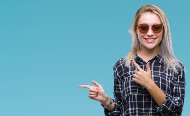 Genç Sarışın kadın gülümseyerek ve iki elleri ve parmakları ile tarafına işaret kamera bakarak izole arka plan üzerinde güneş gözlüğü takıyor.