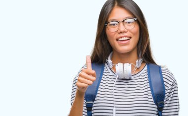 Genç Asyalı öğrenci kadın hareketi el ile mutlu yaşasın yapıyor izole arka plan üzerinde kulaklık ve sırt çantası giyiyor. Kameraya bakarak başarı gösteren ile ifade onaylama.