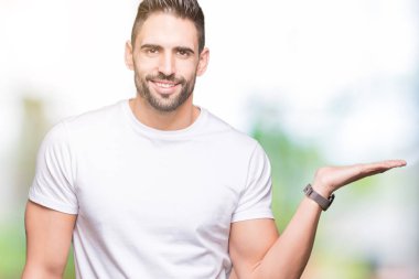 Neşeli sunulması ve kameraya bakarak avucunun ile işaret gülümseyen izole arka plan üzerinde rahat beyaz t-shirt giyen genç adam.
