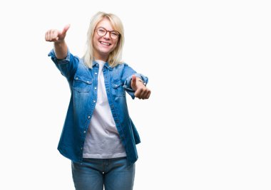 Genç güzel sarışın kadın el ile pozitif jest yapıyor onaylama izole arka plan üzerinde gözlük takan başparmak gülümseyen ve başarı için mutlu. Kamera, kazanan jest arıyorsunuz.
