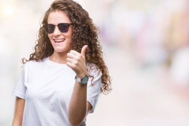 Güzel esmer kıvırcık saçlı Genç kız mutlu yaşasın el ile jest yapıyor izole arka plan üzerinde güneş gözlüğü takıyor. Kameraya bakarak başarı gösteren ile ifade onaylama.