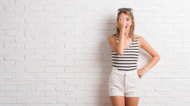 Beyaz tuğla duvar kaplama el yüz ve sürpriz duygu kendinden emin gülümseme ile tek gözlü genç güzel sarışın kadın.