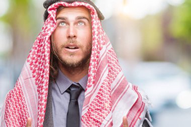 Genç yakışıklı Arap adam uzun saçlı keffiyeh izole arka plan üzerinde giyen hayrete ve yukarıya bakıyor ve parmakları ile işaret ve silah kaldırdı şaşırttı.