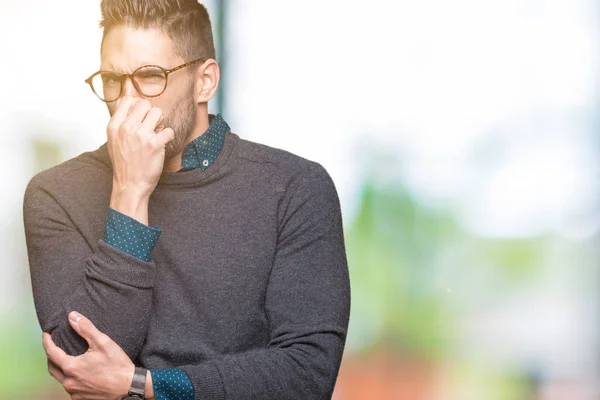 Young handsome man wearing glasses over isolated background smelling something stinky and disgusting, intolerable smell, holding breath with fingers on nose. Bad smells concept.