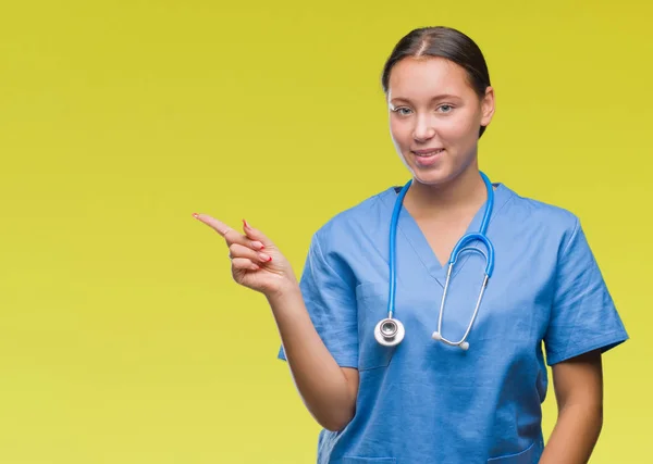 年轻的高加索医生妇女穿着医疗制服在孤立的背景与一个很大的笑容在脸上 用手和手指指着一边看着镜头 — 图库照片