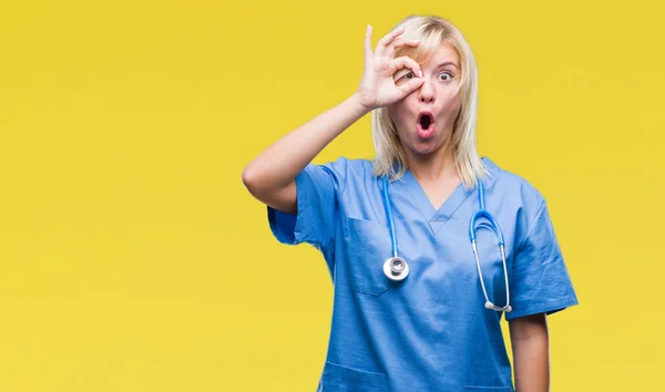 Young beautiful blonde doctor woman wearing medical uniform over isolated background doing ok gesture shocked with surprised face, eye looking through fingers. Unbelieving expression.