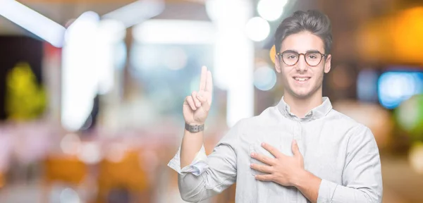 Young handsome man wearing glasses over isolated background Swearing with hand on chest and fingers, making a loyalty promise oath