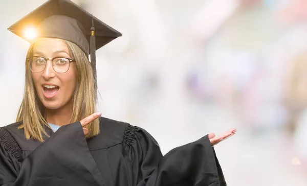 Young beautiful woman wearing graduated uniform over isolated background amazed and smiling to the camera while presenting with hand and pointing with finger.