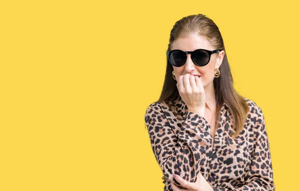 Middle age mature rich woman wearing sunglasses and leopard dress over isolated background looking stressed and nervous with hands on mouth biting nails. Anxiety problem.
