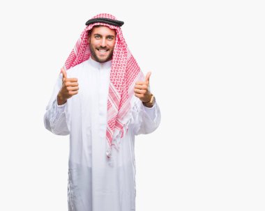 Keffiyeh izole arka plan başarı işareti el ile pozitif jest yapıyor üzerinde giyen genç yakışıklı adam başparmak gülümseyen ve mutlu. Neşeli bir ifadeyle, kazanan jest kameraya bakıyor.