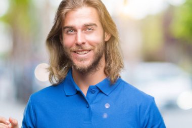 Kameraya bakarak yan el ve parmak ile işaret eden genç yakışıklı adam yüzünde kocaman bir gülümseme ile izole arka plan üzerinde uzun saçlı.