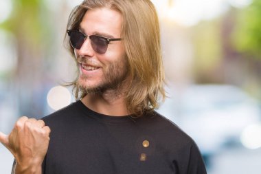 Seyir ve başparmak yanında yukarıyı mutlu yüz ile gülümseyen izole arka plan üzerinde güneş gözlüğü takmış uzun saçlı yakışıklı delikanlı.