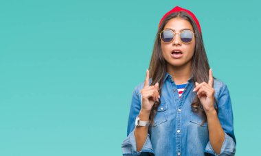 Genç güzel Arap kadın hayrete ve yukarıya bakıyor ve parmakları ile işaret ve silah kaldırdı sürpriz izole arka plan üzerinde güneş gözlüğü takıyor.