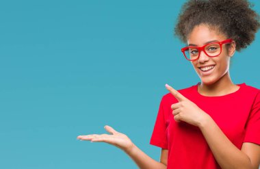 Genç afro Amerikalı kadın hayrete ve el ile sunulması ve parmak ile işaret ederken kameraya gülümseyen izole arka plan üzerinde gözlük.