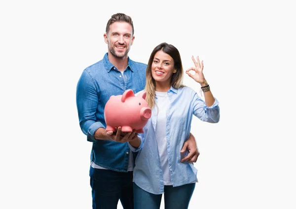 Young couple in love holding piggy bank over isolated background doing ok sign with fingers, excellent symbol