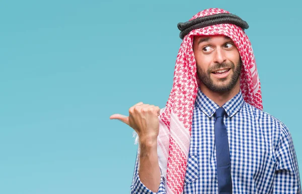 Young handsome arabian business man wearing keffiyeh over isolated background smiling with happy face looking and pointing to the side with thumb up.