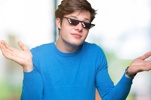 Young man wearing funny thug life glasses over isolated background clueless and confused expression with arms and hands raised. Doubt concept.