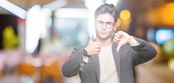 Young business man wearing glasses over isolated background Doing thumbs up and down, disagreement and agreement expression. Crazy conflict