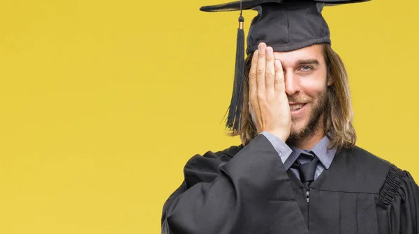 若いハンサムな顔と驚きの感情に自信を持って笑顔で手で片目を覆う孤立の背景の上の長い髪の男の卒業 — ストック写真