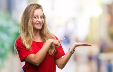 Genç güzel sarışın kadın hayrete ve el ile sunulması ve parmak ile işaret ederken kameraya gülümseyen izole arka plan üzerinde gözlük.