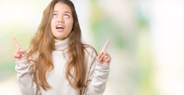 Hayret ve yukarıya bakıyor ve parmakları ile işaret ve silah kaldırdı sürpriz izole arka plan üzerinde boğazlı kazak giyen genç güzel esmer kadın.