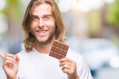 Uzun saç beslenme ile yakışıklı delikanlı çikolata bar üzerinde izole arka tarafına el ve parmak ile işaret çok mutlu