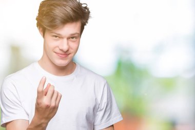 Beckoning gel burada mutlu ve gülümseyerek el davet ile jest izole arka plan üzerinde rahat beyaz t-shirt giyen genç yakışıklı adam