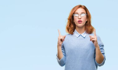 İzole arka plan kış kazak giyen genç güzel kadın hayrete ve yukarıya bakıyor ve parmakları ile işaret ve silah kaldırdı şaşırttı.