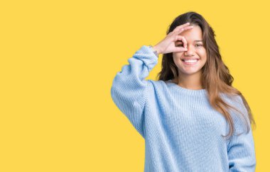 Genç güzel esmer kadın giyiyor mavi kış kazak gülümseyen, parmakları ile mutlu yüz üzerinden seyir göz el ile ok jest yapıyor izole arka plan üzerinde.