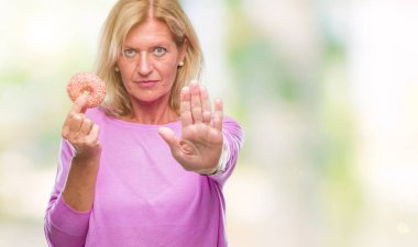 Orta yaş sarışın kadın ile izole arka plan üzerinde pembe çörek yeme dur işareti ile ciddi ve kendine güvenen ifade, savunma hareketi yaparken el açmak