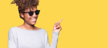 Kameraya bakarak yan el ve parmak ile işaret yüzünde kocaman bir gülümseme ile izole arka plan üzerinde güneş gözlüğü takıyor güzel genç Afro-Amerikan kadın.