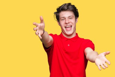 Sarılmak için kollarını açarak gülümseyerek kameraya bakarak izole arka plan üzerinde kırmızı t-shirt giyen genç yakışıklı adam. Neşeli ifade kucaklayan mutluluk.