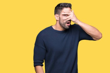 Mahcup ifadeyle parmaklarımın arasından seyir yüz ve gözler el ile kapsayan şok bakmak izole arka plan üzerinde kazak giyen genç yakışıklı adam.