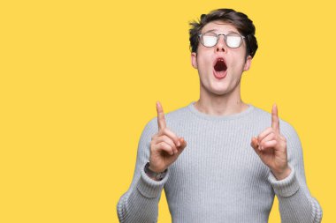 Yakışıklı delikanlı hayrete ve yukarıya bakıyor ve parmakları ile işaret ve silah kaldırdı sürpriz izole arka plan üzerinde gözlük.