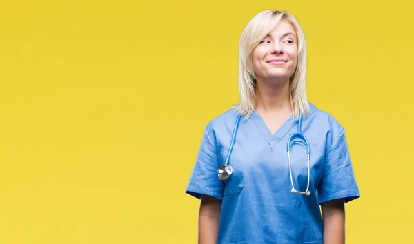 Young beautiful blonde doctor woman wearing medical uniform over isolated background smiling looking side and staring away thinking.