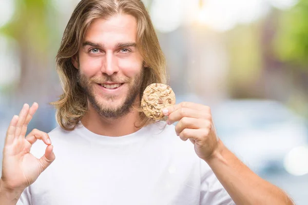 優秀なシンボルの指で サインをして孤立した背景にチョコレート クッキーを食べて髪の長い若いハンサムな男 — ストック写真
