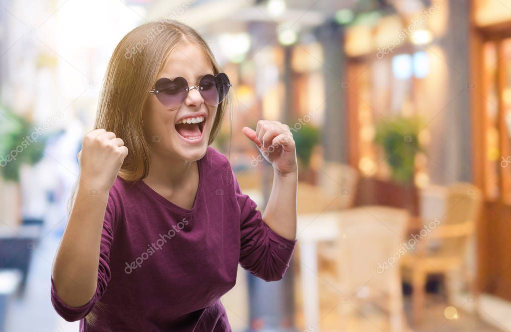 Young beautiful girl wearing sunglasses over isolated background very happy and excited doing winner gesture with arms raised, smiling and screaming for success. Celebration concept.
