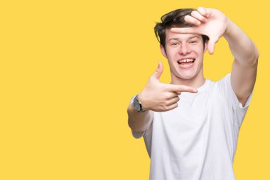 Elle yapma çerçeve ve parmakları ile mutlu yüz gülümseyen izole arka plan üzerinde rahat beyaz t-shirt giyen genç yakışıklı adam. Yaratıcılık ve Fotoğrafçılık kavramı.
