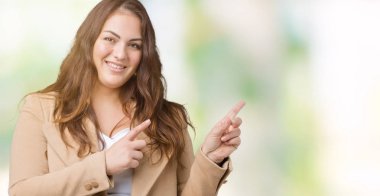 Artı boyutu genç kadın güzel üzerinde kış palto giyiyor gülümseyen ve iki elleri ve parmakları ile tarafına işaret kamera bakarak arka plan izole.