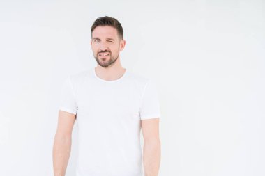 Seksi bir ifadeyle, neşeli ve mutlu yüzünü kameraya bakarak göz kırpıyor izole arka plan üzerinde rahat beyaz t-shirt giyen genç yakışıklı adam.