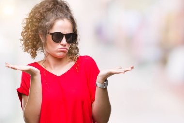 Güzel esmer kıvırcık saçlı Genç kız üzerinde güneş gözlüğü takıyor arka plan clueless ve şaşkın bir ifadeyle kollar ve eller kaldırdı izole. Şüphe kavramı.