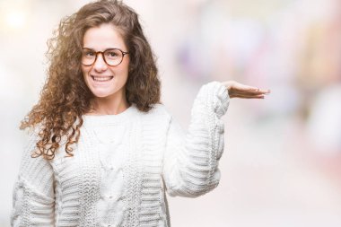 Neşeli sunulması ve kameraya bakarak avucunun ile işaret gülümseyen izole arka plan üzerinde kış kazak giyen güzel esmer kıvırcık saçlı Genç kız.