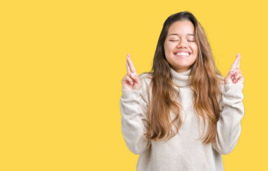 Umut ve gözleri kapalı geçiş parmaklarıyla gülümseyen izole arka plan üzerinde boğazlı kazak giyen genç güzel esmer kadın. Şans ve batıl kavramı.