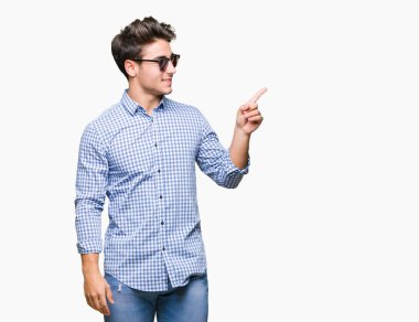 Kameraya bakarak yan el ve parmak ile işaret eden genç yakışıklı adam yüzünde kocaman bir gülümseme ile izole arka plan üzerinde güneş gözlüğü takıyor.