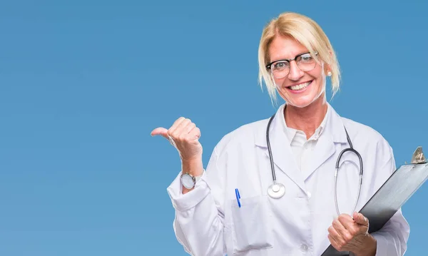 中年金发碧眼的医生妇女拿着剪贴板在孤立的背景指向和显示与快乐的脸微笑的一侧 — 图库照片