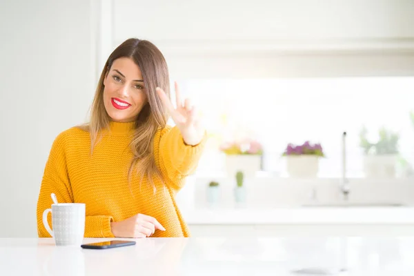 Young beautiful woman drinking a cup of coffee at home smiling looking to the camera showing fingers doing victory sign. Number two.