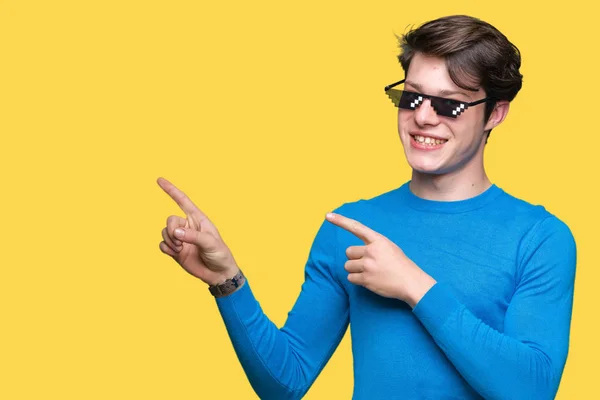 Young man wearing funny thug life glasses over isolated background smiling and looking at the camera pointing with two hands and fingers to the side.