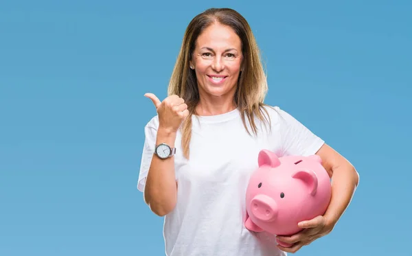 中年西班牙妇女存钱使用存钱罐在孤立的背景指向和显示与幸福的脸微笑的一侧 — 图库照片