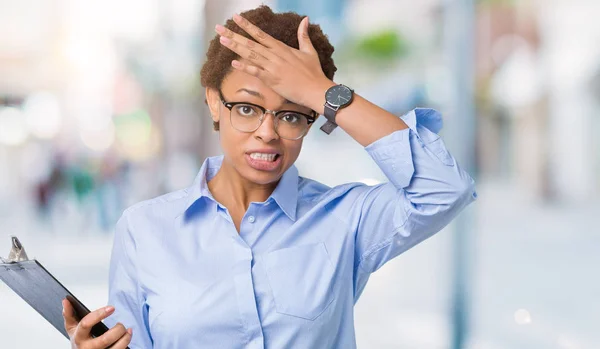 Young african american businesss woman holding clipboard over isolated background stressed with hand on head, shocked with shame and surprise face, angry and frustrated. Fear and upset for mistake.